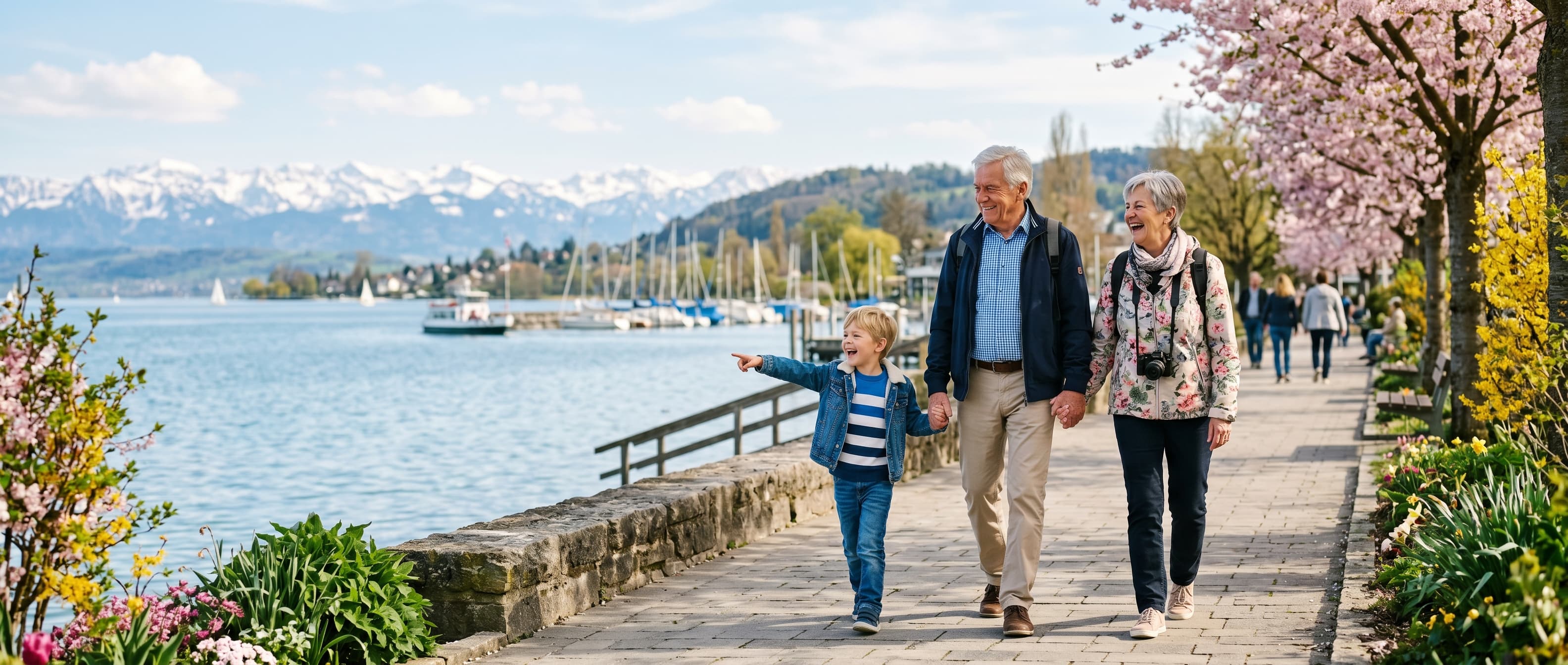 Großeltern mit Enkel auf einer Uferpromenade am Bodensee, Frühlingsblüte und Alpenpanorama im Hintergrund